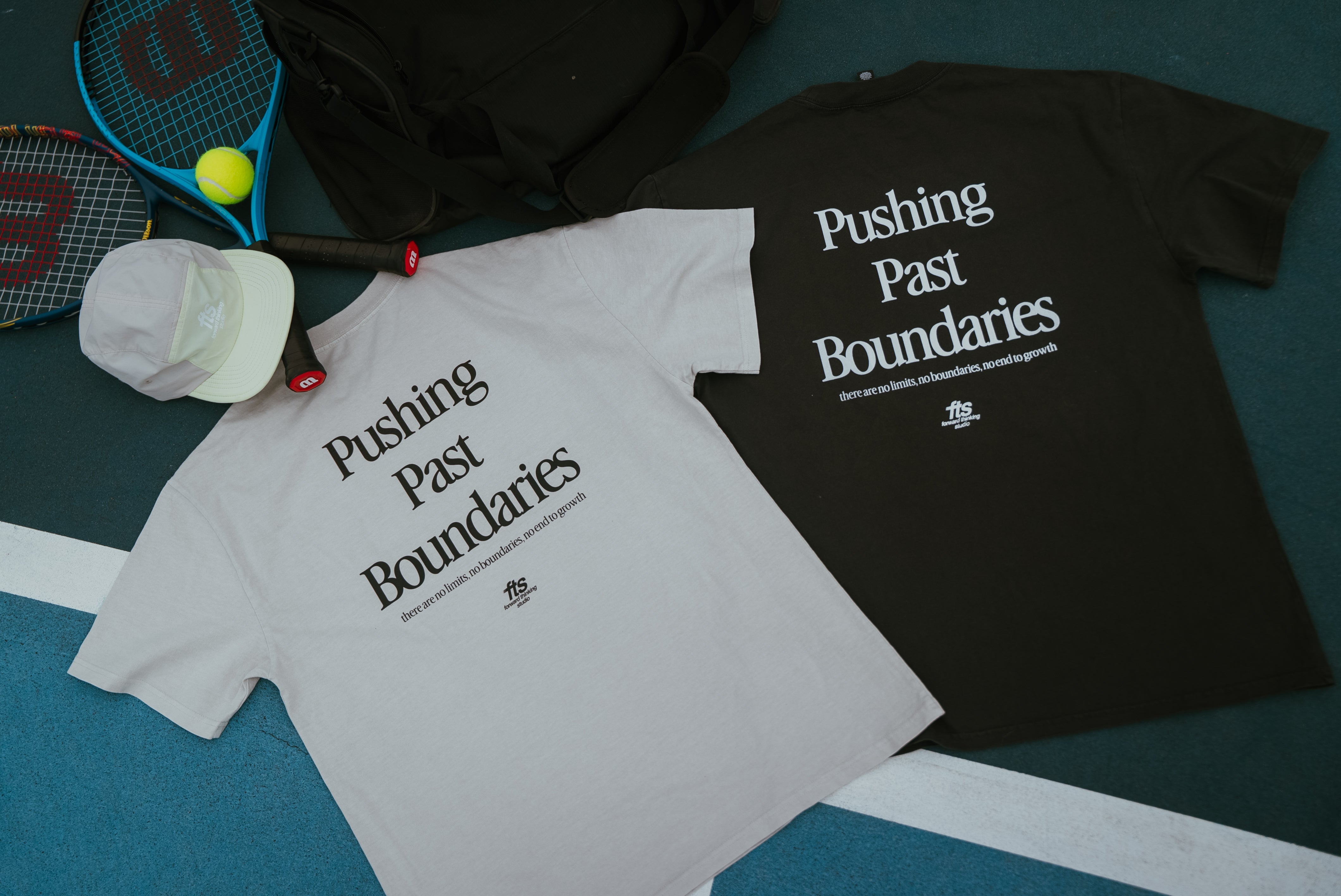 Two FTS t-shirts with 'Pushing Past Boundaries' text on a sports-themed surface with tennis rackets and balls.